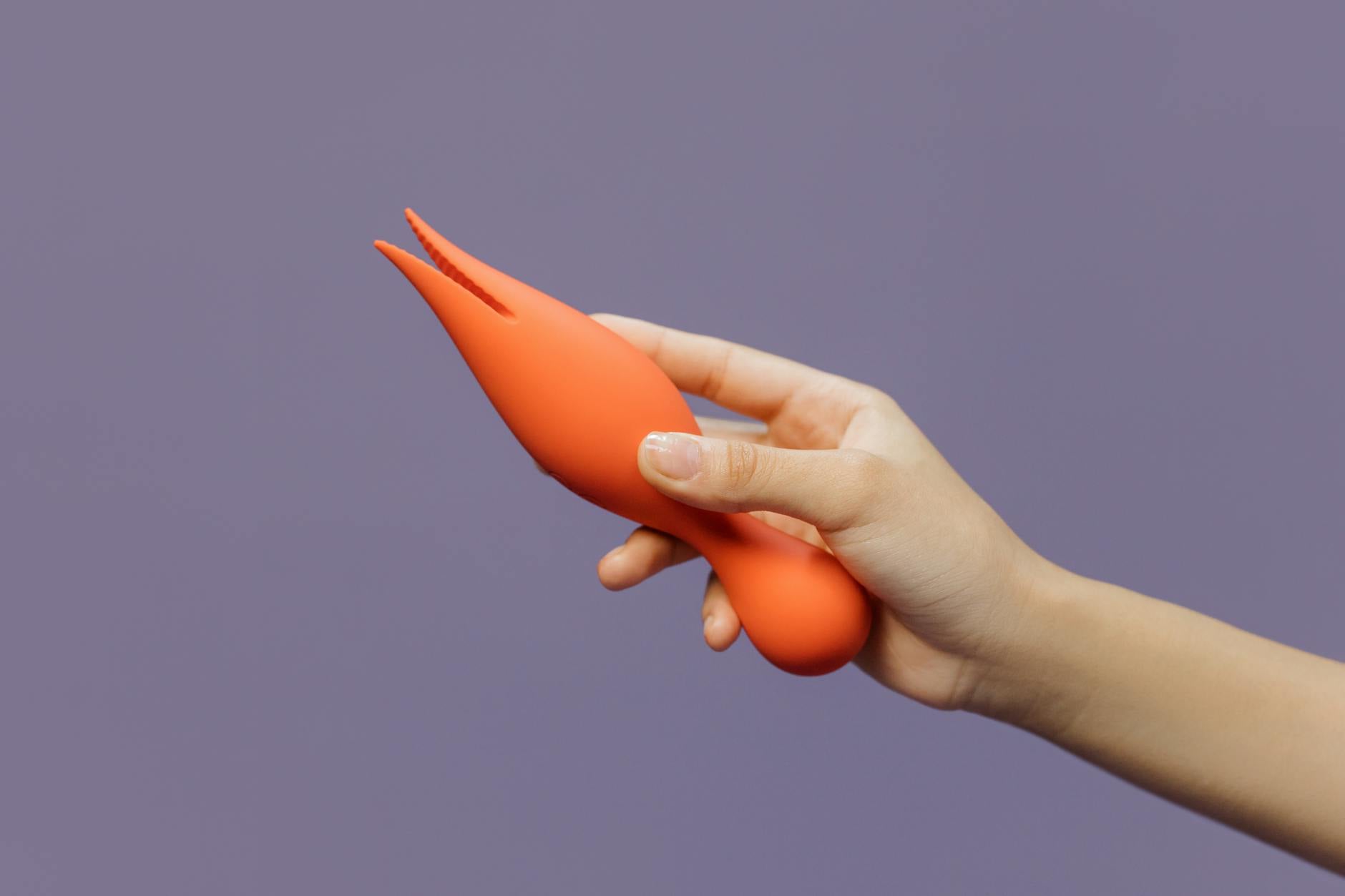 Two hands holding pink and blue silicone vibrators against a pastel background